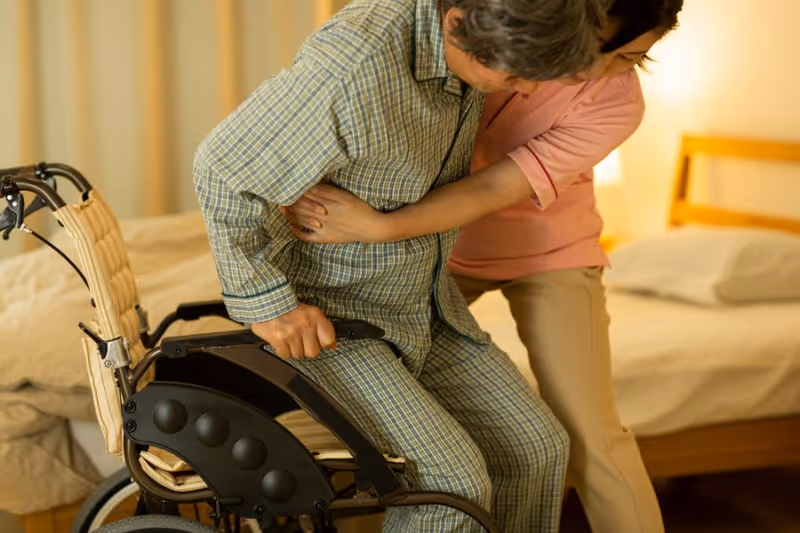 A caregiver assists an elderly person in pajamas to stand up from a wheelchair in a bedroom setting with a bed and soft lighting in the background.