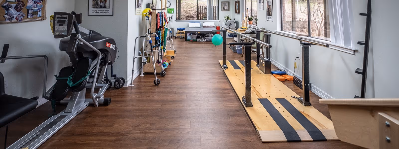 A rehabilitation or physical therapy room with exercise equipment including a SciFit machine, parallel bars for walking practice, walkers, resistance bands, and therapy balls. The room has wooden flooring, large windows on the right side, and a bench with pillows at the far end.
