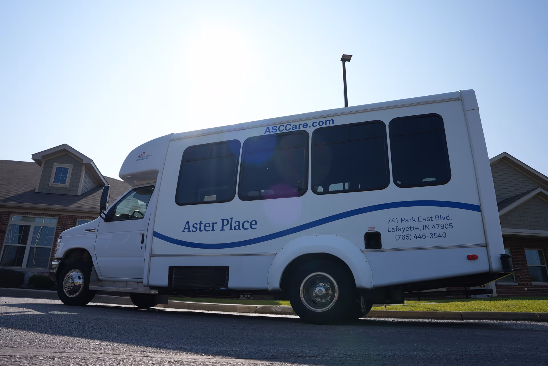 A white shuttle bus parked on a street in front of residential-style buildings. The bus has the name 'Aster Place' written on its side along with the website ASCCare.com and the address 741 Park East Blvd., Lafayette, IN 47905, and phone number (765) 446-3540.