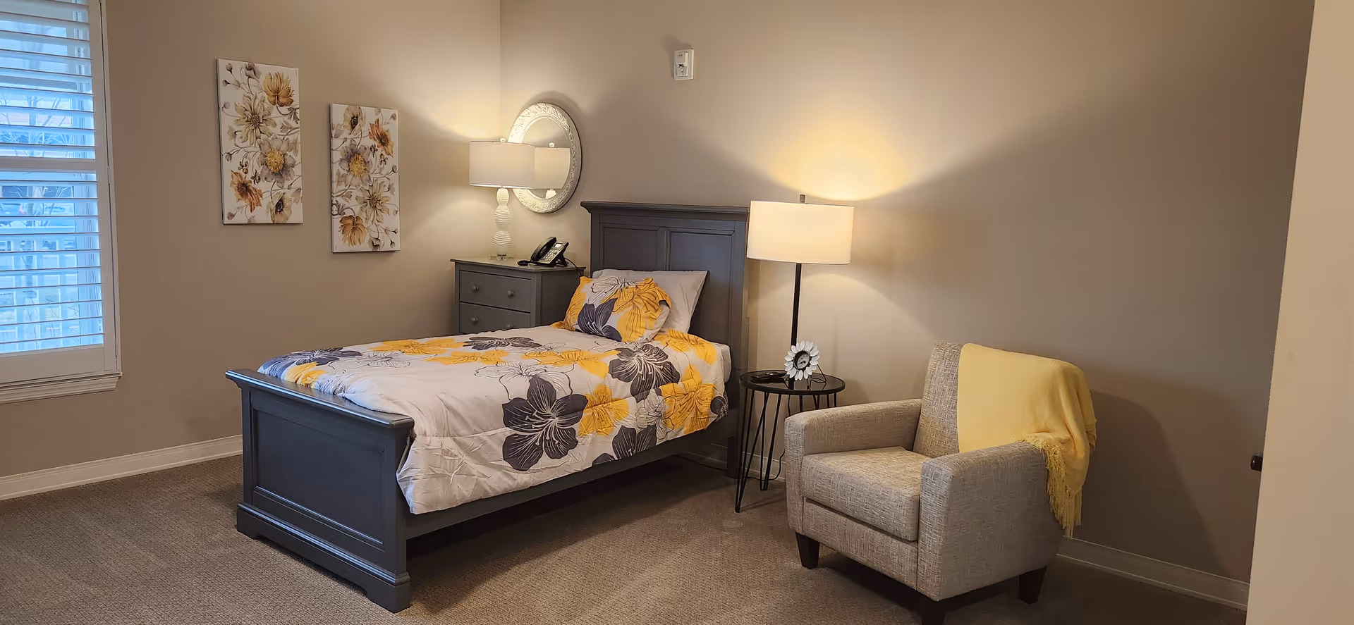A cozy bedroom with a single bed featuring a floral yellow and gray comforter. Next to the bed is a gray nightstand with a lamp, a phone, and a round mirror above it. A beige armchair with a yellow throw blanket is placed beside a small round black side table with a lamp and a framed photo. Two floral paintings hang on the wall near a window with white blinds.