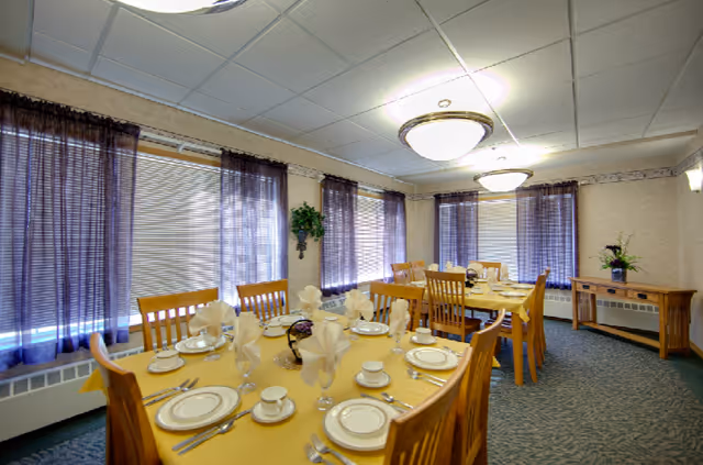 A dining room with two wooden tables set with white china plates, cups, saucers, silverware, and folded napkins. The room has several windows with purple curtains, carpeted floor, beige walls, ceiling lights, and a wooden side table with a flower arrangement.