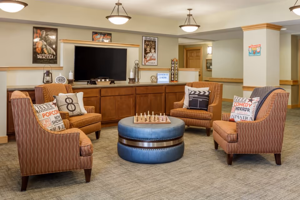 Cozy lounge area with four upholstered armchairs arranged around a round ottoman and a TV on a wooden cabinet.
