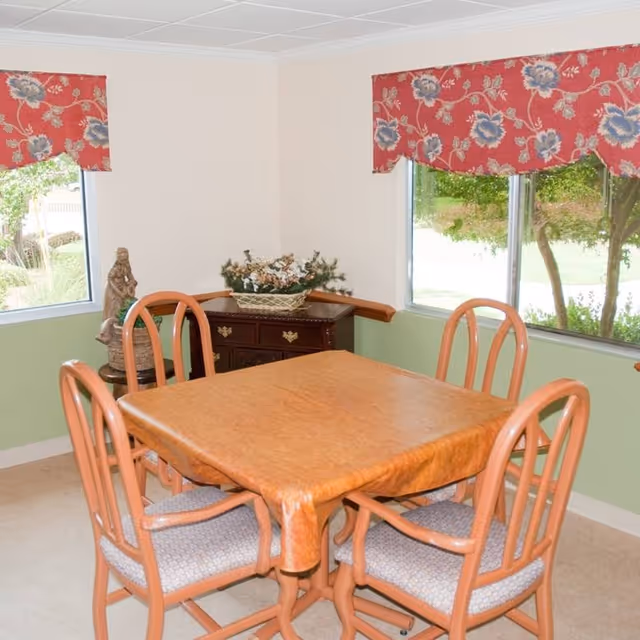 A small dining area with a square wooden table covered by a brown tablecloth and four wooden chairs with cushioned seats. The room has two windows with red floral valances, light green lower walls, and cream upper walls. A dark wooden cabinet with a floral arrangement and a small statue is placed in the corner between the windows.