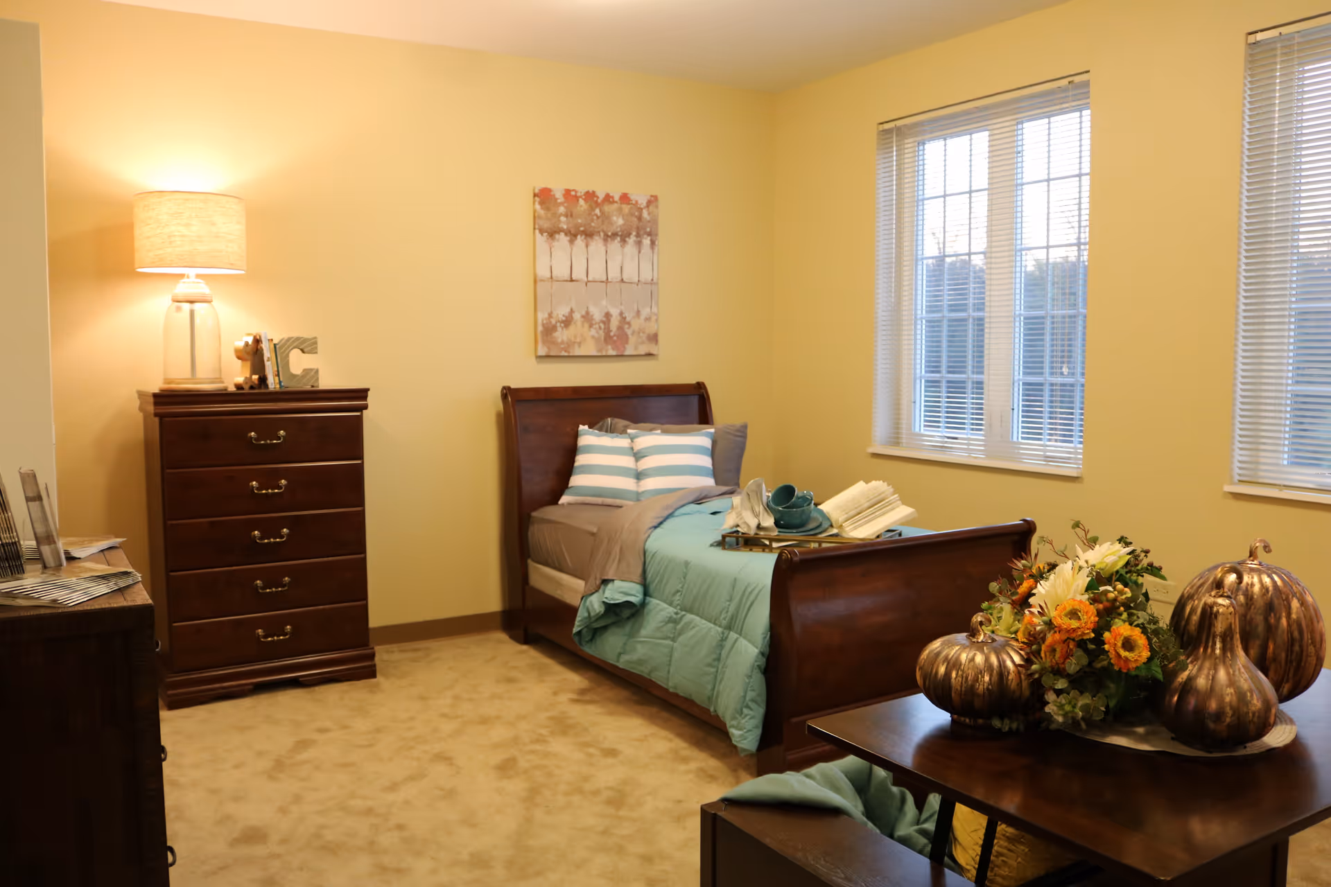 Furnished bedroom with a wooden sleigh bed dressed in teal bedding, a tall dresser with a lamp, windows, and a table topped with floral and pumpkin decorations.