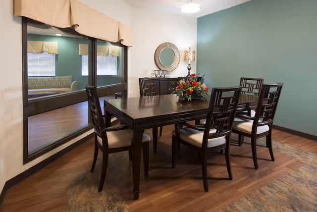 A dining room with a dark wooden table and six matching chairs with cream cushions. There is a floral centerpiece on the table. The room has a teal accent wall with a round mirror and a lamp on a sideboard. A large window with beige valances looks into another room with a couch.