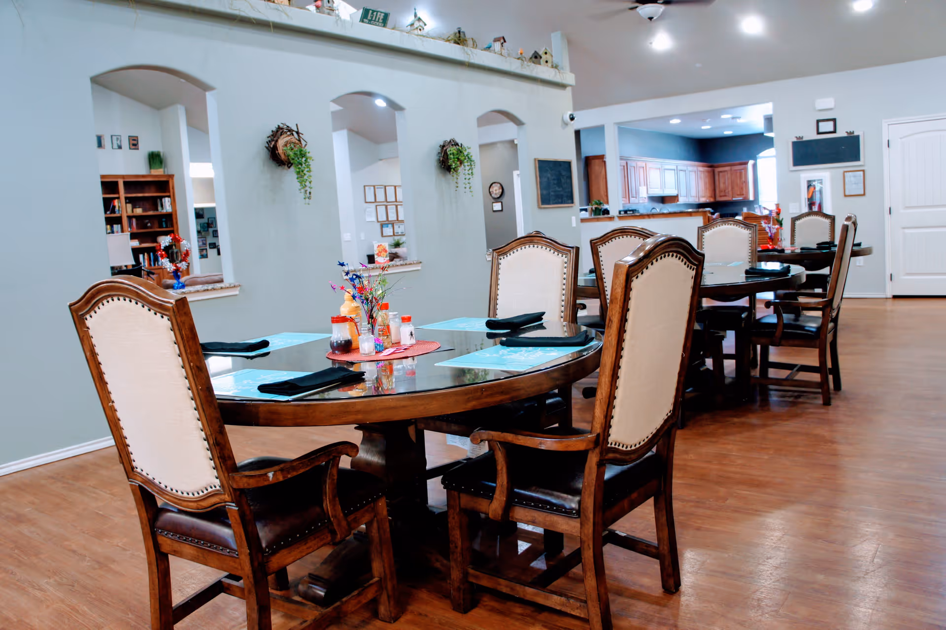 Interior view of a senior living facility dining area with wooden tables and cushioned chairs. The tables are set with placemats, napkins, and condiments. The room has hardwood flooring, light-colored walls, and an open layout with a kitchen visible in the background. Decorative plants hang on the wall arches.