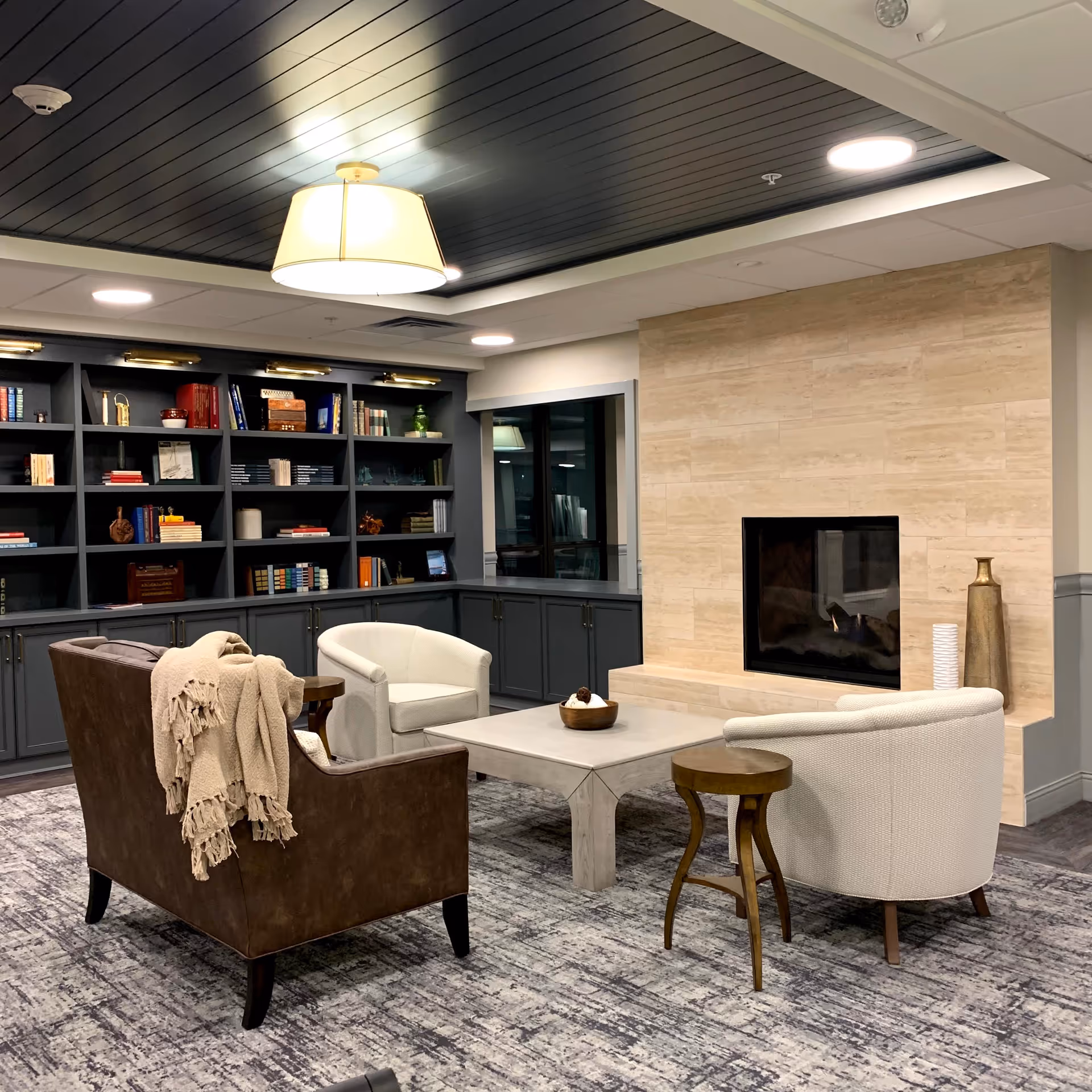 A cozy living room area with a brown leather loveseat draped with a beige throw blanket, two white upholstered armchairs, a square coffee table, and a small wooden side table. The room features a modern fireplace with a beige stone surround, built-in dark gray bookshelves filled with books and decorative items, and a dark wood paneled ceiling with a large hanging light fixture.