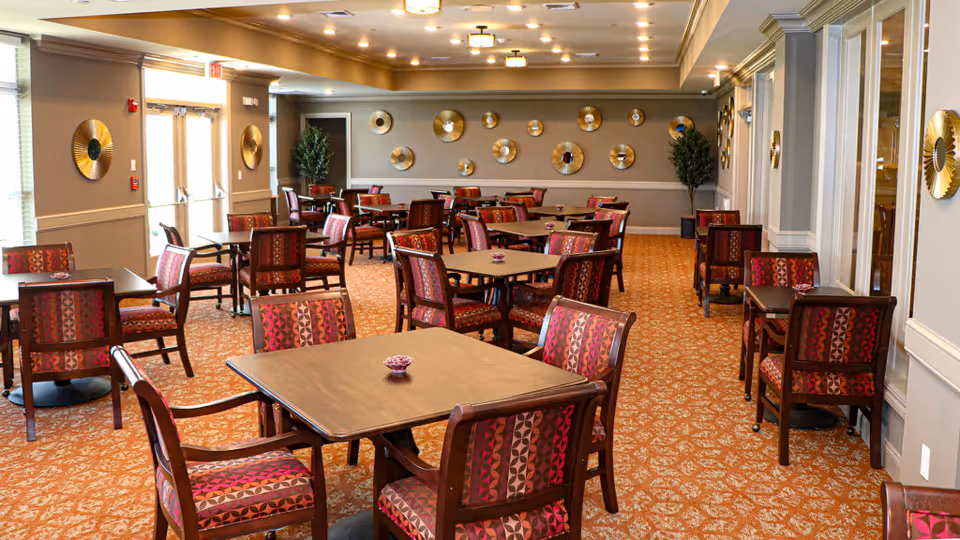 A spacious dining room with multiple wooden tables and chairs featuring red and purple patterned upholstery. The room has beige walls decorated with circular gold wall art and two potted plants in the corners. The floor is carpeted with a patterned design, and there are large windows letting in natural light.