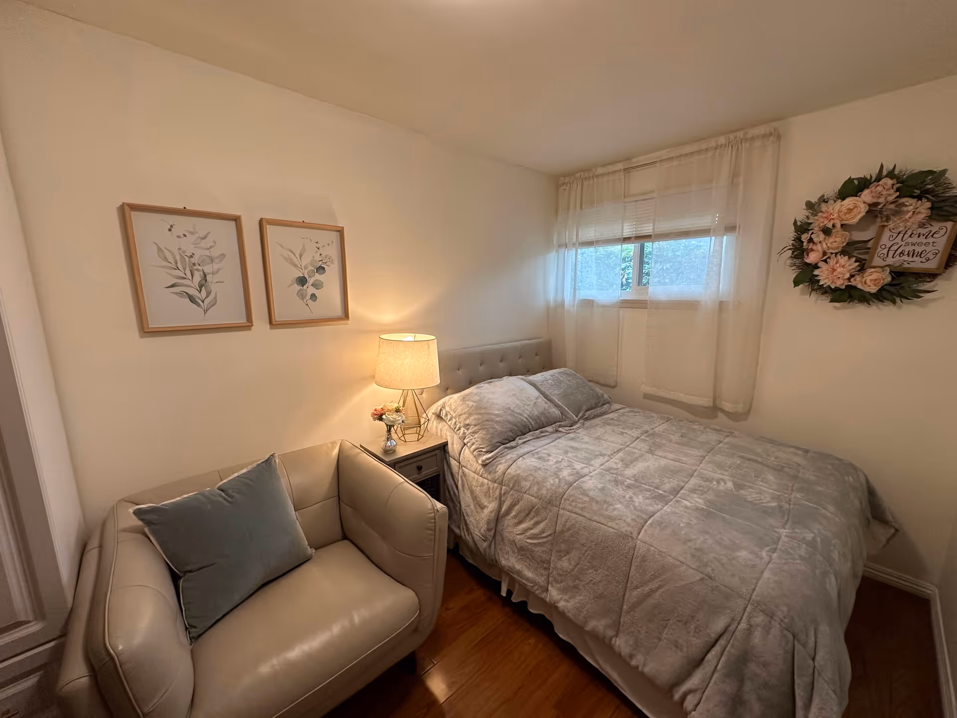 Small cozy bedroom with a bed, nightstand and lamp, an armchair, framed botanical prints and a decorative wreath on the wall.