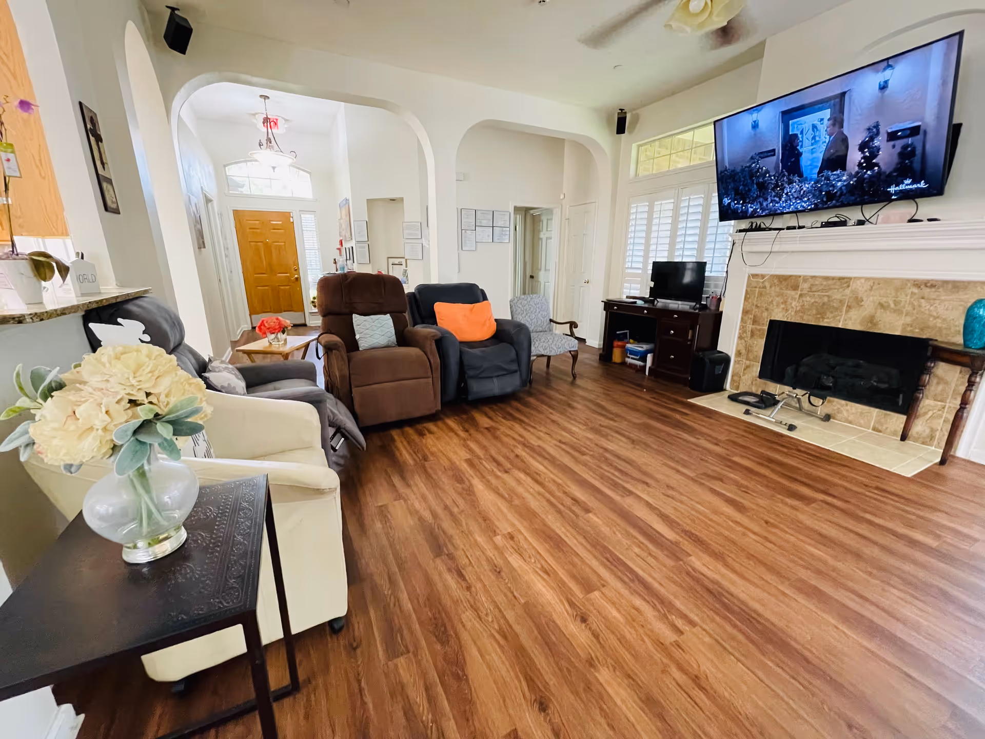 A cozy living room with wooden flooring, several armchairs including a white chair with a side table holding a vase of flowers, a brown recliner, a dark gray recliner with an orange pillow, and a patterned armchair. A large flat-screen TV is mounted above a tiled fireplace, and there is a small desk with a computer monitor in the corner. The room has white walls, arched doorways, and a wooden front door visible in the background.