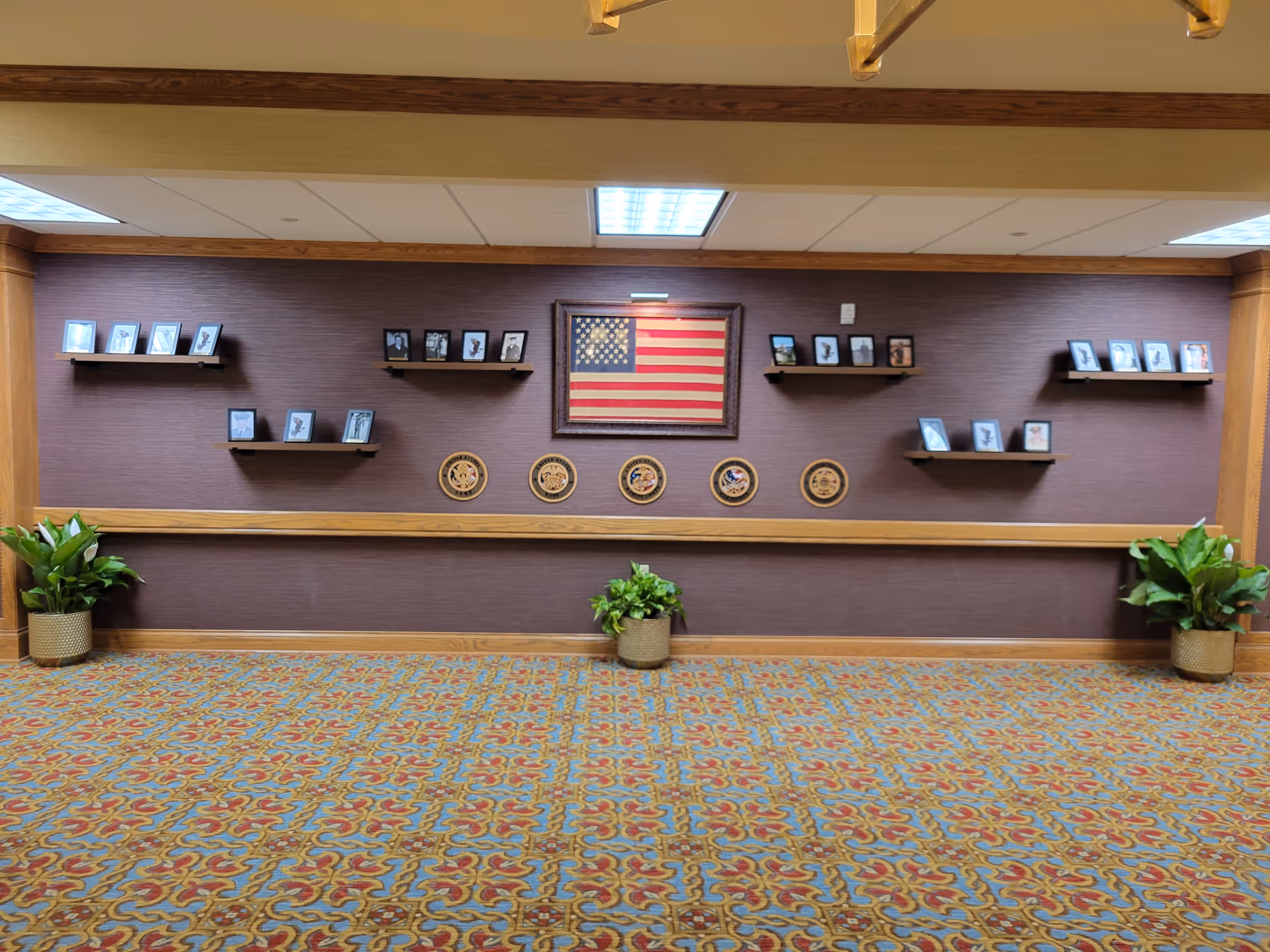 Interior hallway wall decorated with framed photographs on small shelves, a framed American flag, and five military service emblems. The wall is brown with wooden trim and has three potted plants on the floor. The carpet has a colorful, intricate pattern.