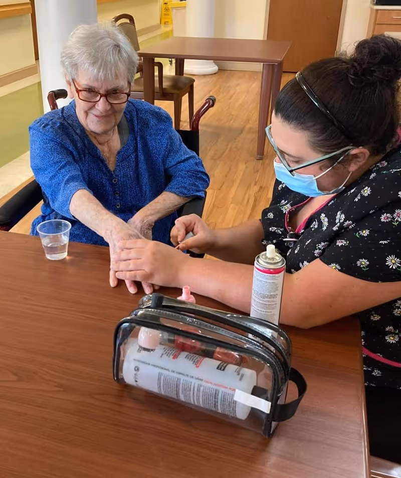 An elderly woman in a blue shirt sitting at a wooden table while a caregiver wearing a mask and floral scrubs paints her nails. On the table, there is a clear bag containing nail care products and a small plastic cup with water.
