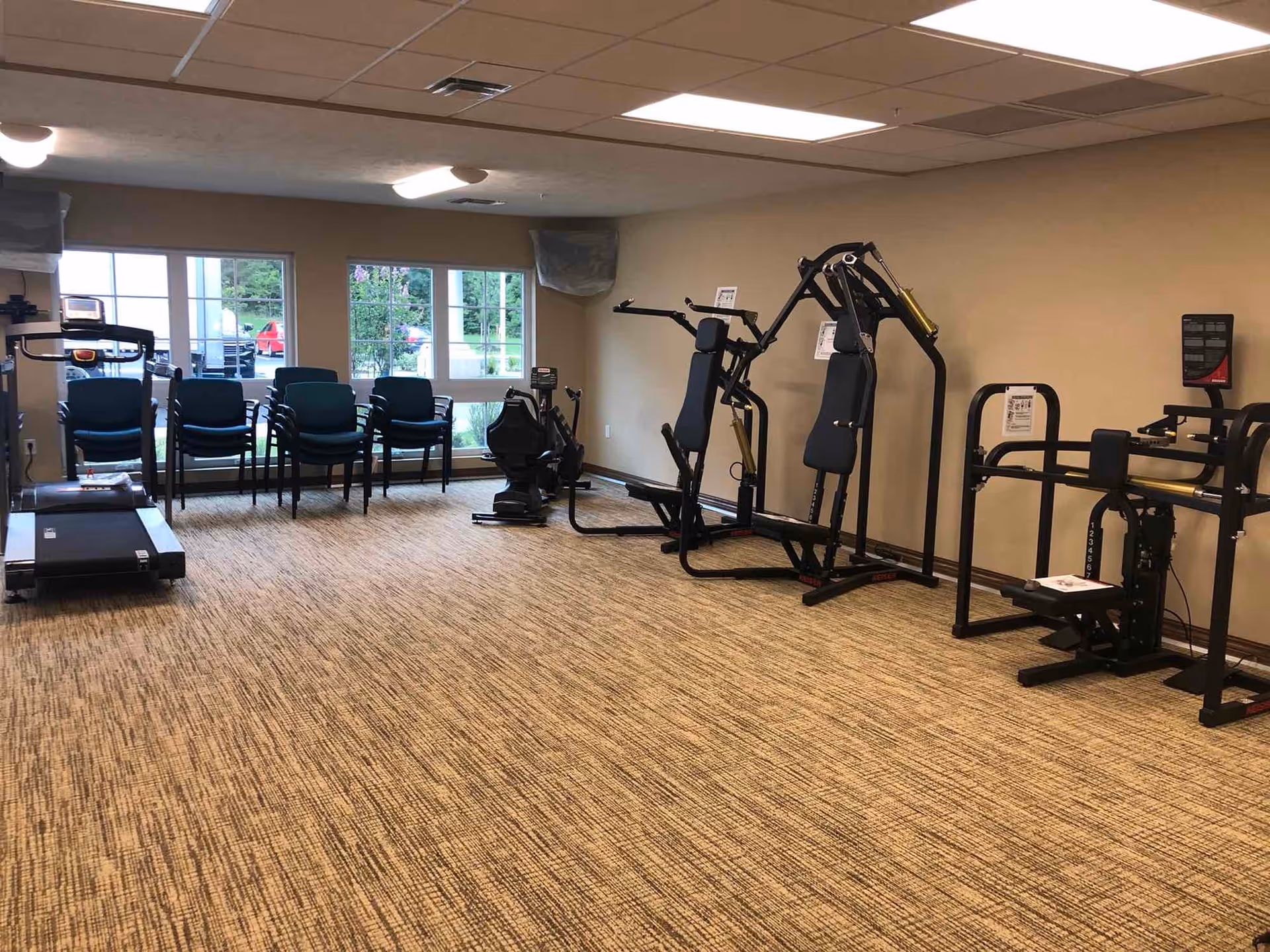 An interior fitness room with a treadmill, weight machines, and stacked chairs in front of windows.