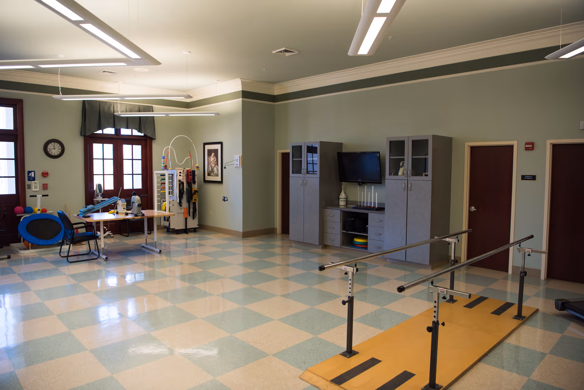 A spacious rehabilitation room with light green walls and checkered light blue and beige flooring. The room contains parallel bars for walking exercises, a table with chairs, exercise equipment, cabinets with a TV mounted on the wall, and large windows with dark wood frames and green valances.