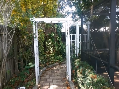 A garden pathway with a white wooden arbor surrounded by greenery and plants. The path is paved with bricks and curves slightly. On the right side, there is a screened enclosure with plants growing nearby. Trees and bushes provide shade and a natural setting.