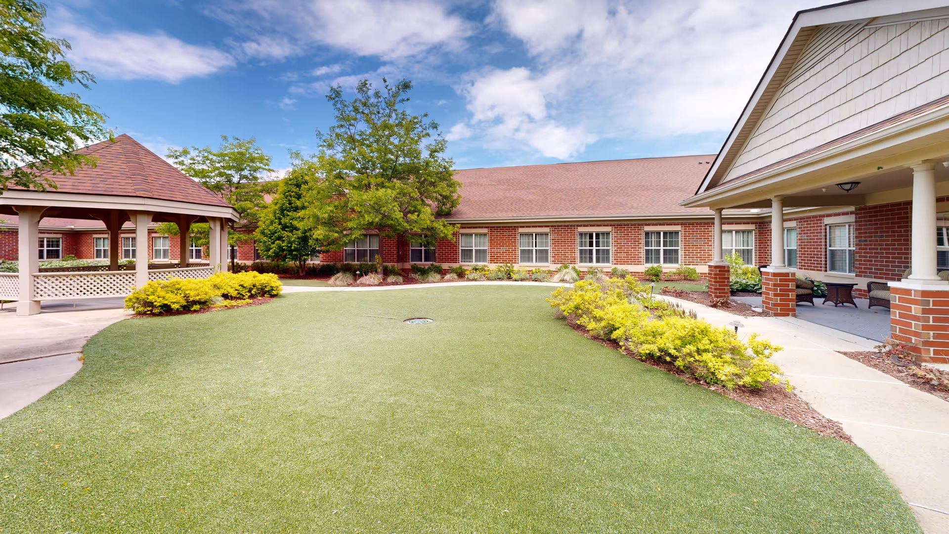 Landscaped courtyard with a gazebo, artificial lawn, walkways and a single-story red-brick assisted living building under a partly cloudy sky.