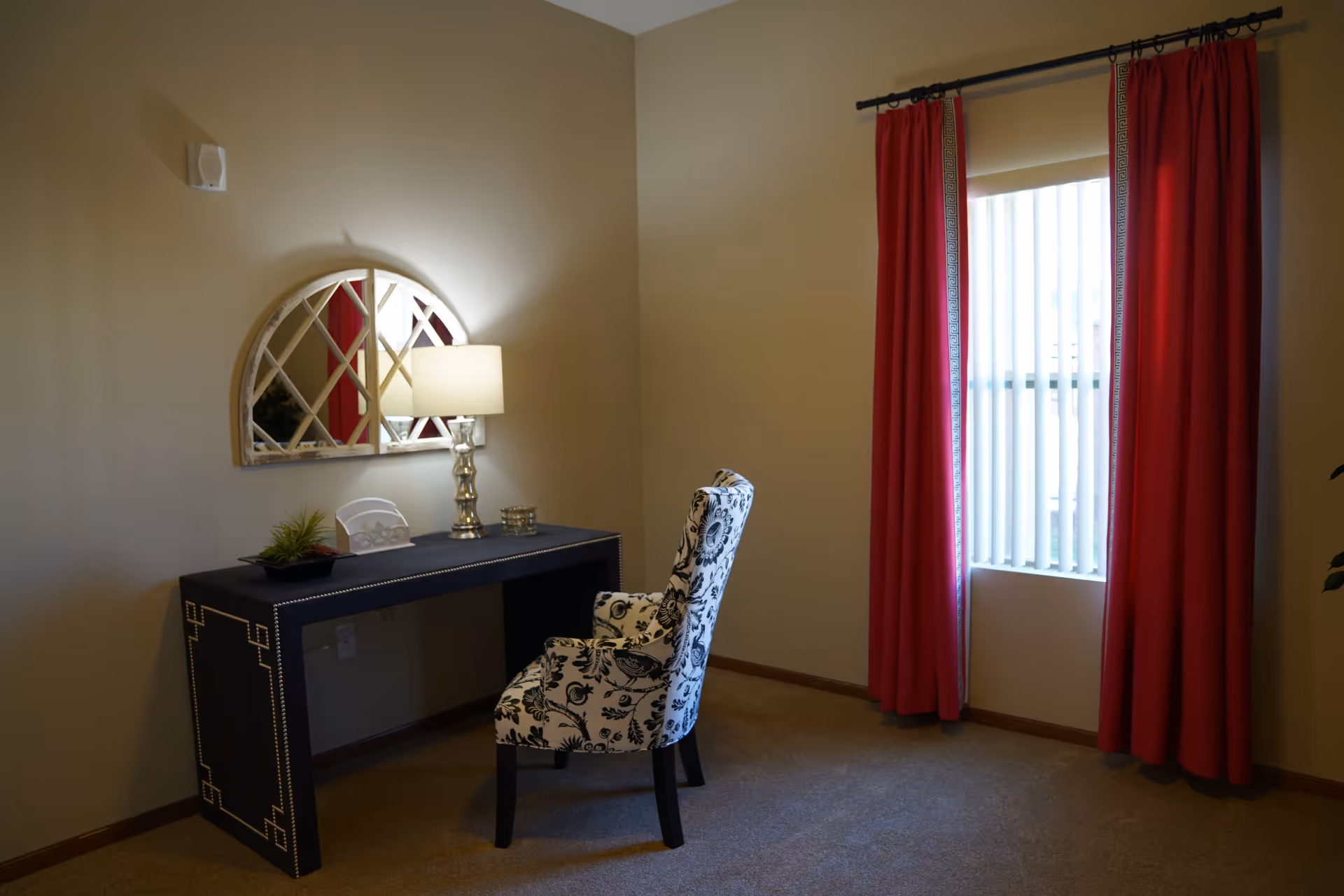 A small room with beige walls and carpeted floor featuring a black desk with decorative nailhead trim, a patterned upholstered chair, a table lamp, a small plant, and a decorative mirror on the wall. There is a window with vertical blinds and red curtains.