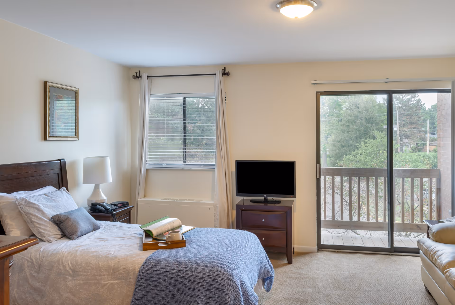 Bedroom with a neatly made bed, bedside table and lamp, a small TV on a dresser, and a sliding glass door leading to a balcony.