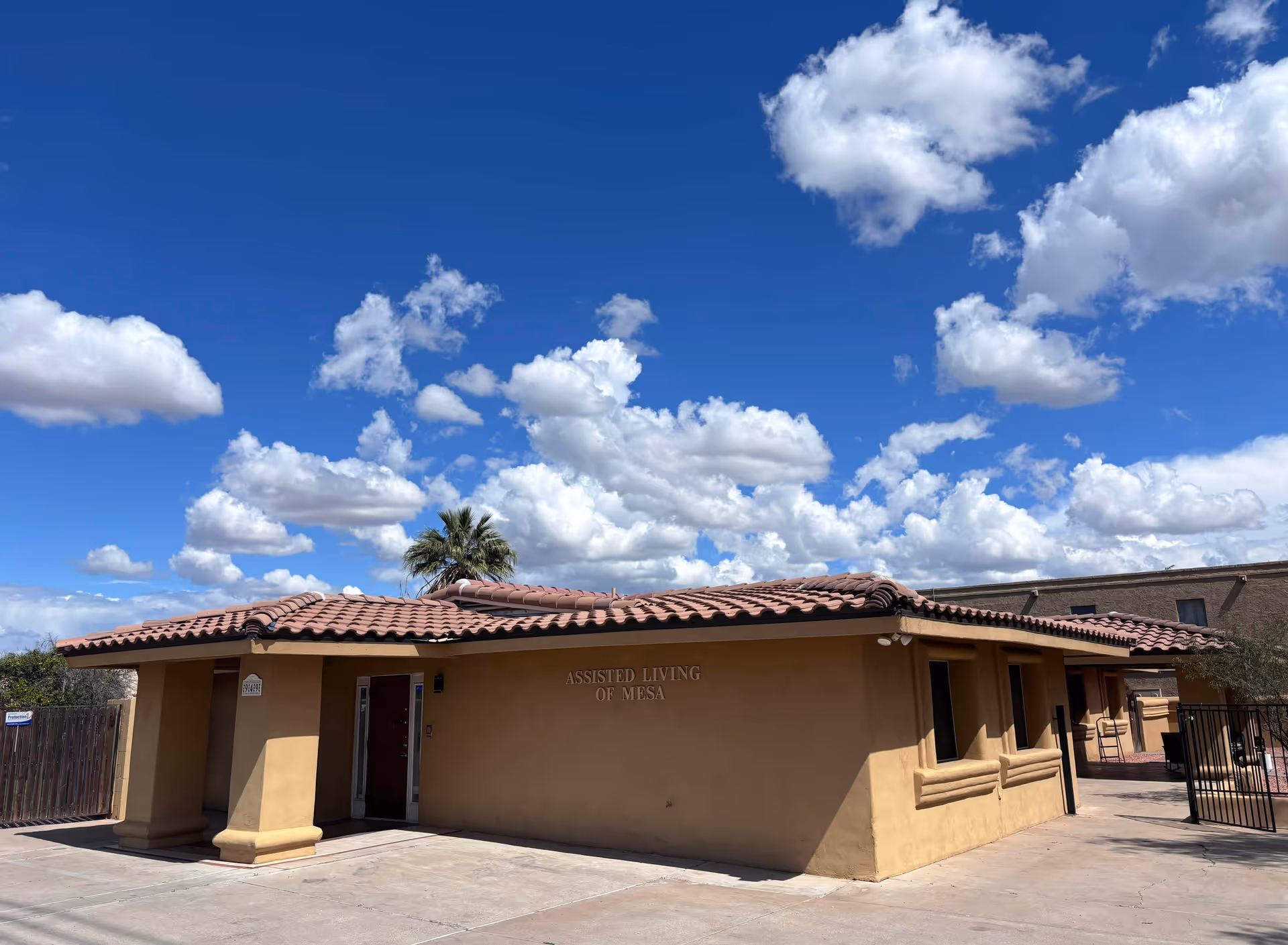 Exterior view of a single-story building with a tan stucco finish and red tile roof under a blue sky with scattered white clouds. The building has a sign that reads 'Assisted Living of Mesa'. There is a palm tree visible behind the building and a paved area in front.