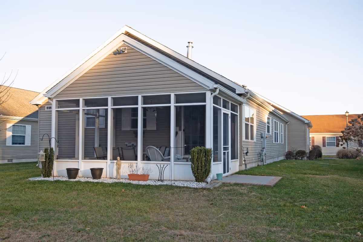 Exterior view of a single-story residential building with beige siding and a screened-in porch. The porch has white framing and contains outdoor furniture. The building is surrounded by a grassy lawn with some small plants and potted flowers near the porch.