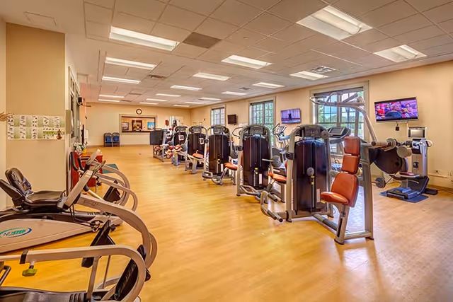 Interior view of a fitness center with multiple exercise machines arranged in rows on a wooden floor. The room is well-lit with ceiling lights and has large windows on one side. There are televisions mounted on the walls and a reception area visible in the background.