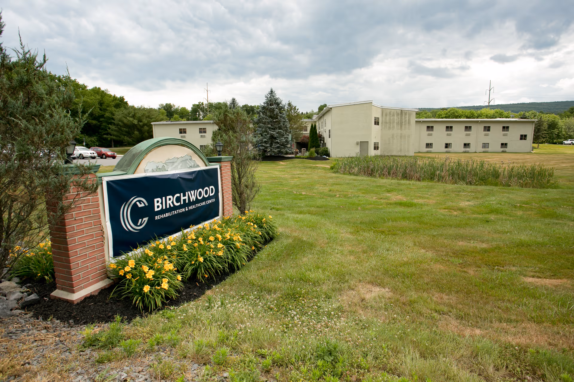 Outdoor view of Birchwood Rehabilitation and Healthcare Center showing a large green lawn with yellow flowers in front of a brick sign displaying the facility's name. The building is visible in the background under a cloudy sky.