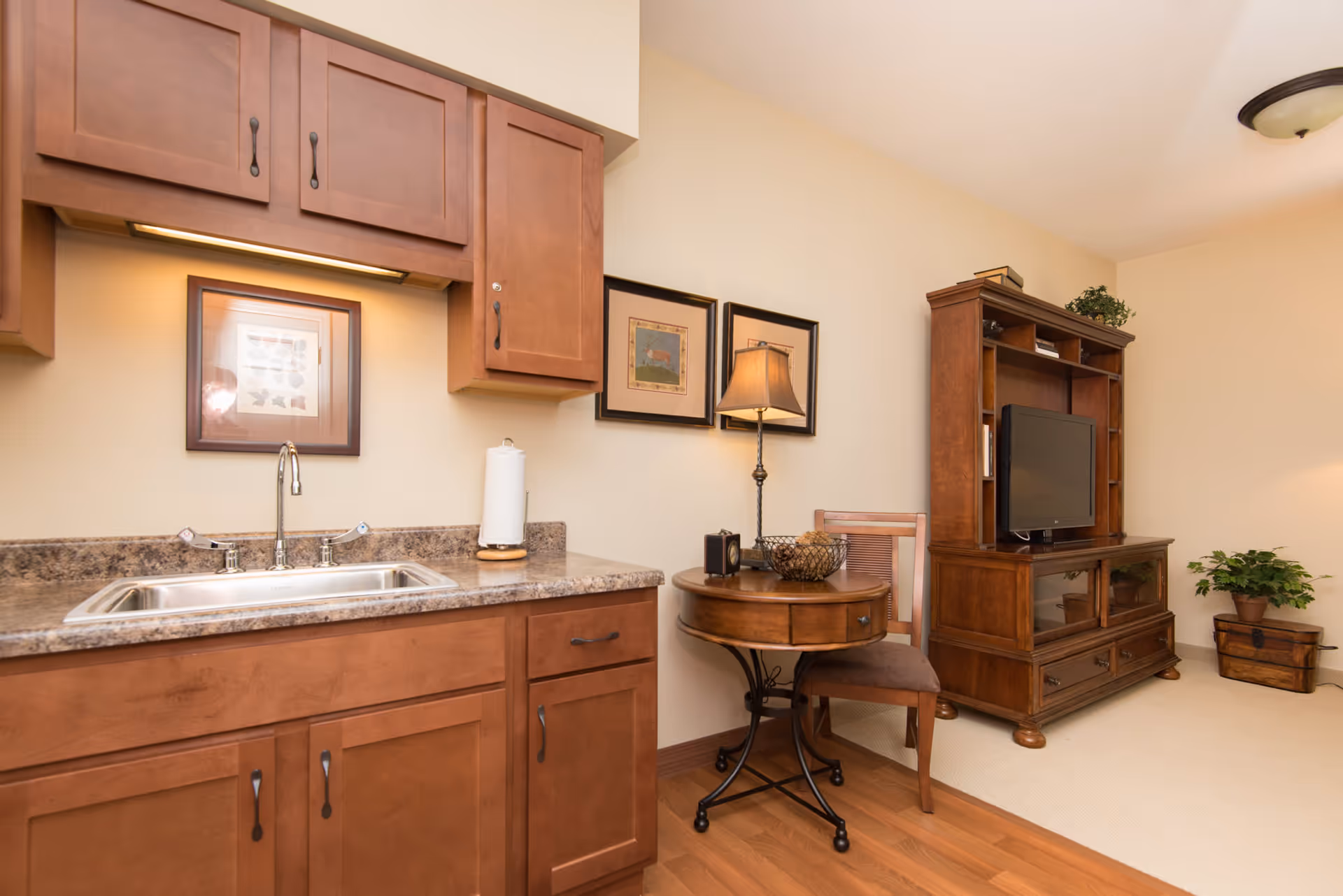 Interior view of a senior living facility room featuring a small kitchenette with wooden cabinets, a sink, and a paper towel holder. Adjacent to the kitchenette is a round wooden table with a lamp, decorative bowl, and a chair. In the background, there is a wooden entertainment center with a flat-screen TV, some books, and a potted plant on the floor next to a wooden chest.
