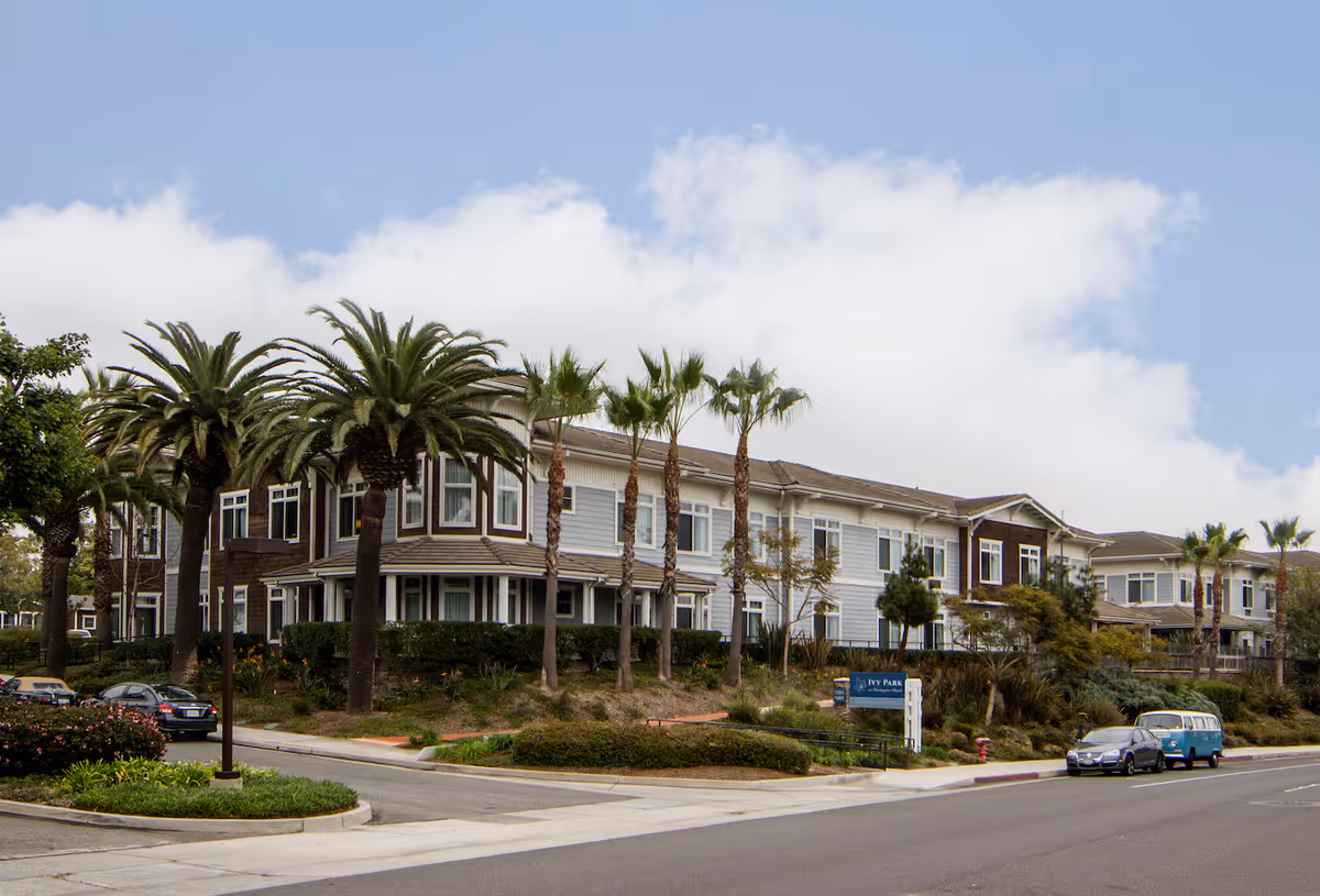 Two-story senior living building with palm trees lining the front and cars parked along the street.