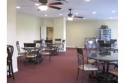 Large common dining area with multiple round glass-topped tables and metal chairs, ceiling fans, maroon carpet and pale green walls.