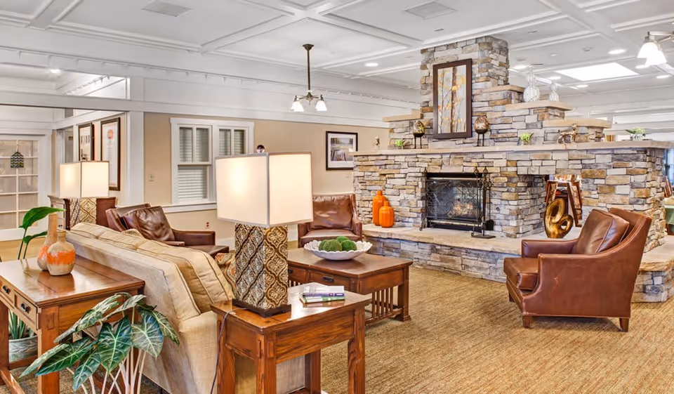 A cozy living room area in a senior living facility featuring a large stone fireplace with decorative items and artwork above it. The room is furnished with brown leather chairs, a beige sofa, wooden side tables with lamps, and plants. The ceiling has white paneling and recessed lighting.