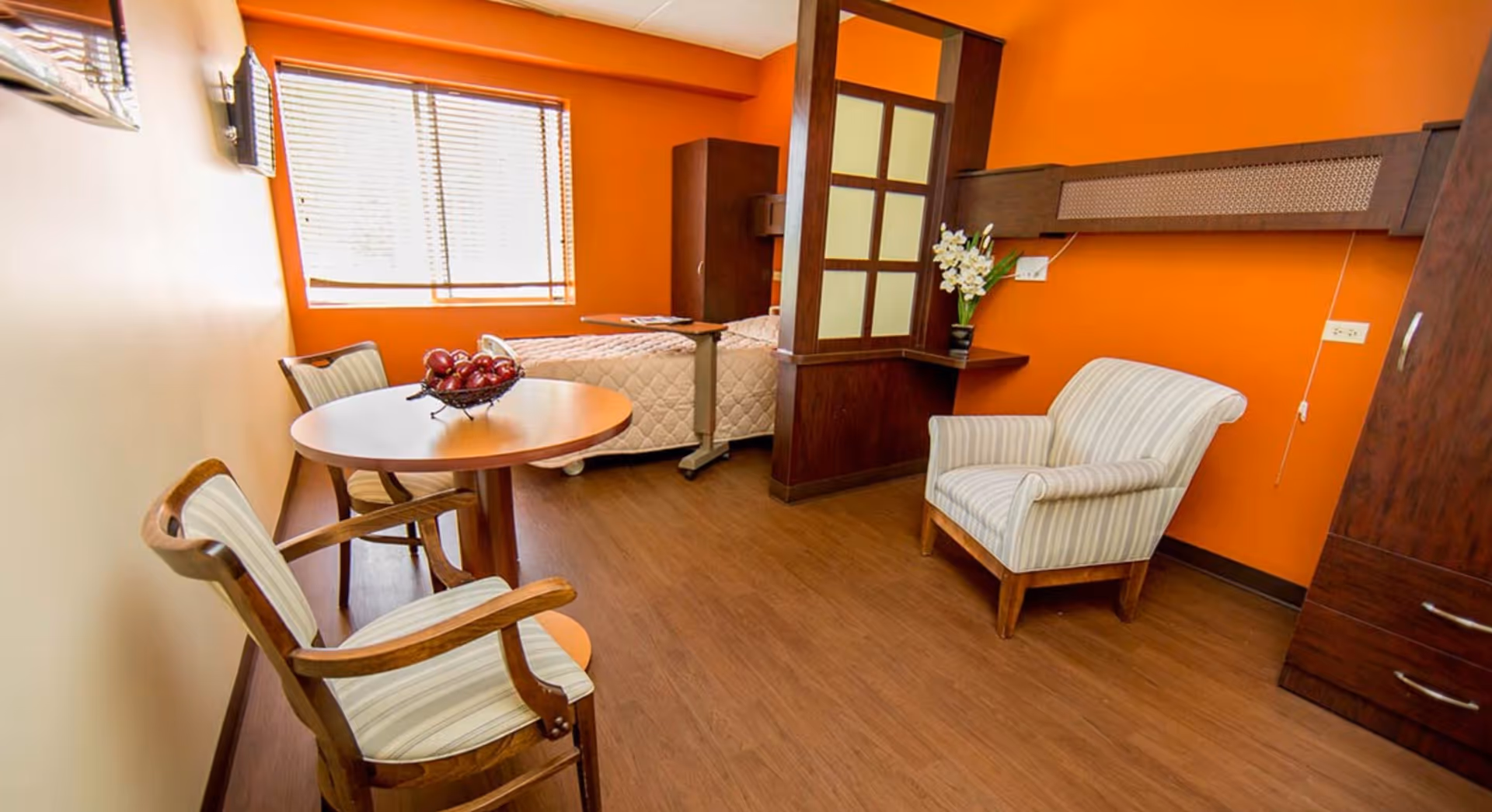 Private senior living studio with an orange accent wall, a bed, round table with chairs, and an upholstered armchair.