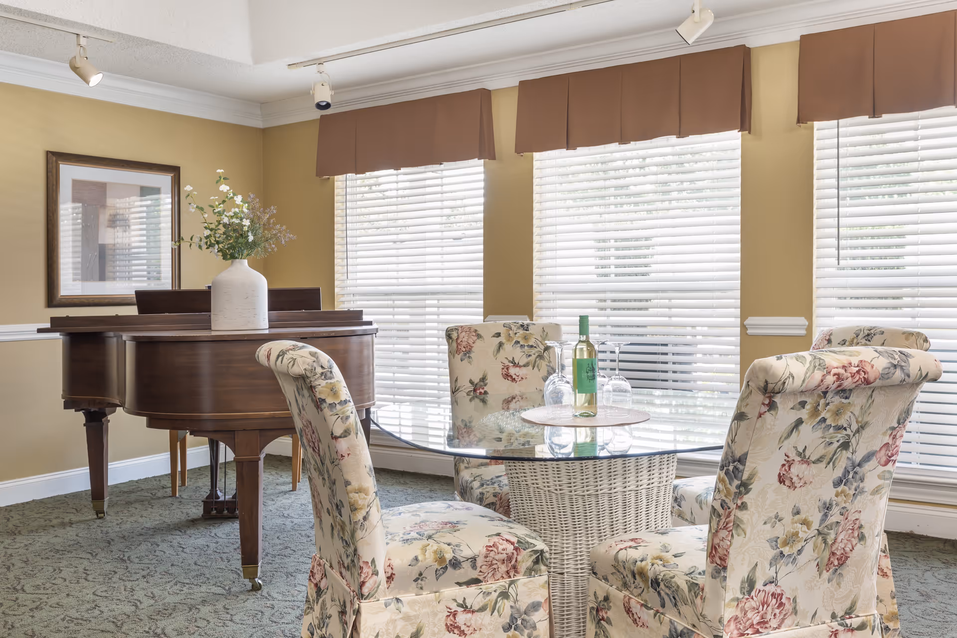 Bright common room with floral upholstered chairs around a glass-top table and a grand piano by large windows.