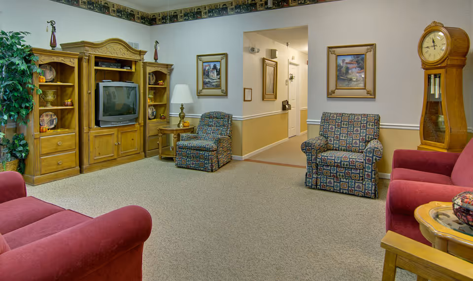 Common sitting room with red sofas, patterned armchairs, a wooden entertainment center holding a TV, a grandfather clock, and a hallway entrance.