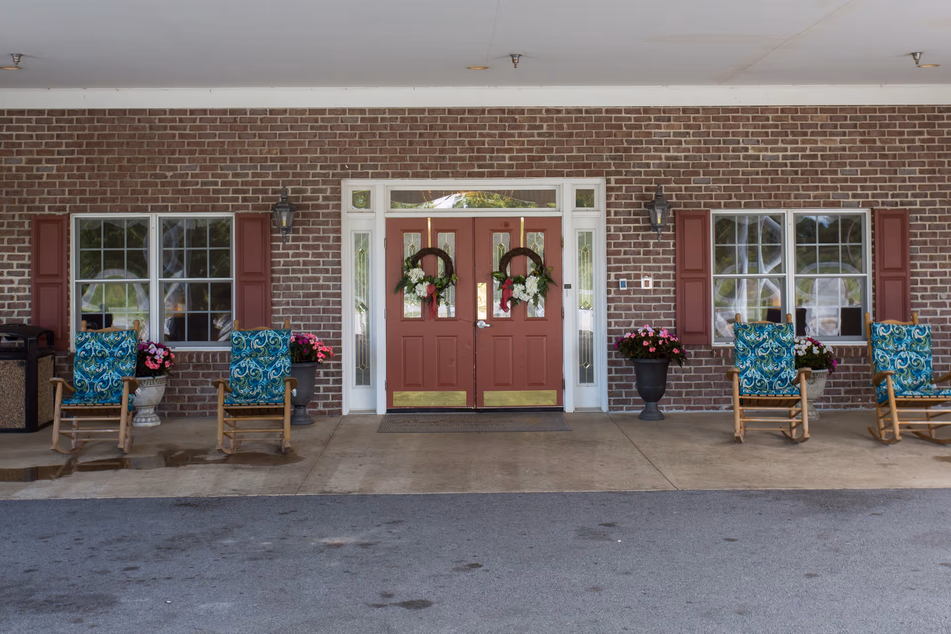 Covered front entrance of a brick assisted living building with double red doors decorated with wreaths, rocking chairs with patterned cushions, and potted flowers.