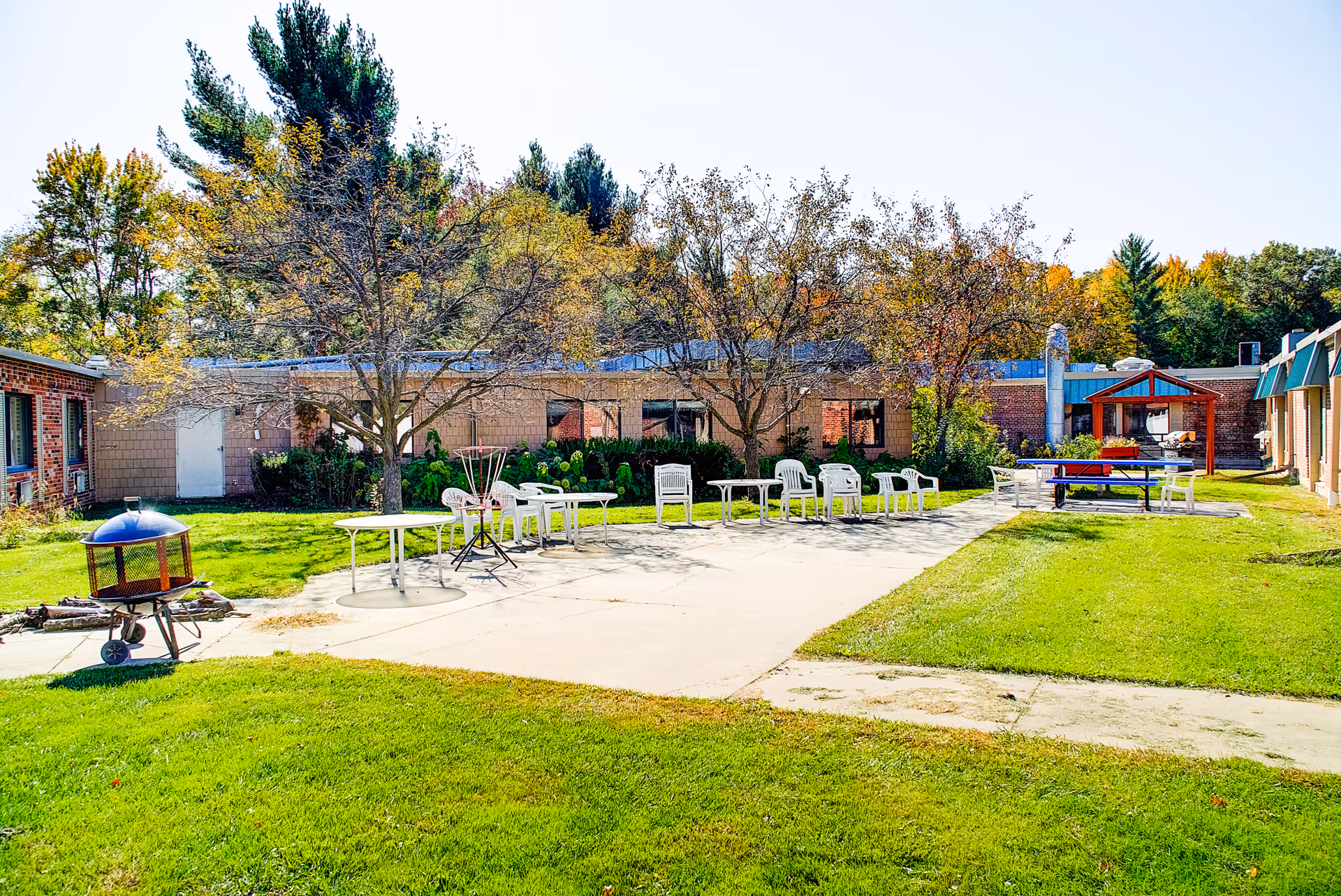 Outdoor courtyard area at Edenbrook Wisconsin Rapids with green grass, trees with autumn leaves, several white plastic chairs and tables arranged on a concrete patio, a fire pit on wheels, and a covered seating area in the background.