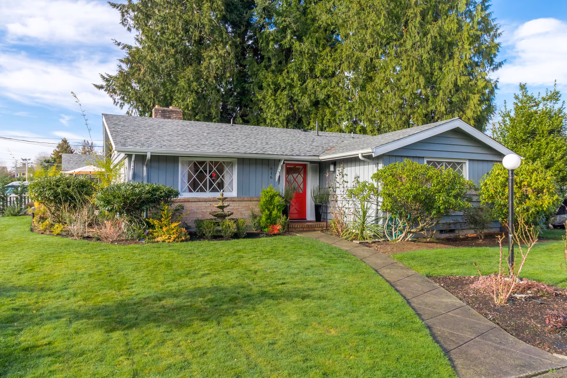Single-story house with blue siding and a red front door, surrounded by a well-maintained green lawn and various shrubs and plants. A curved concrete pathway leads to the entrance, and tall trees are visible in the background under a partly cloudy sky.