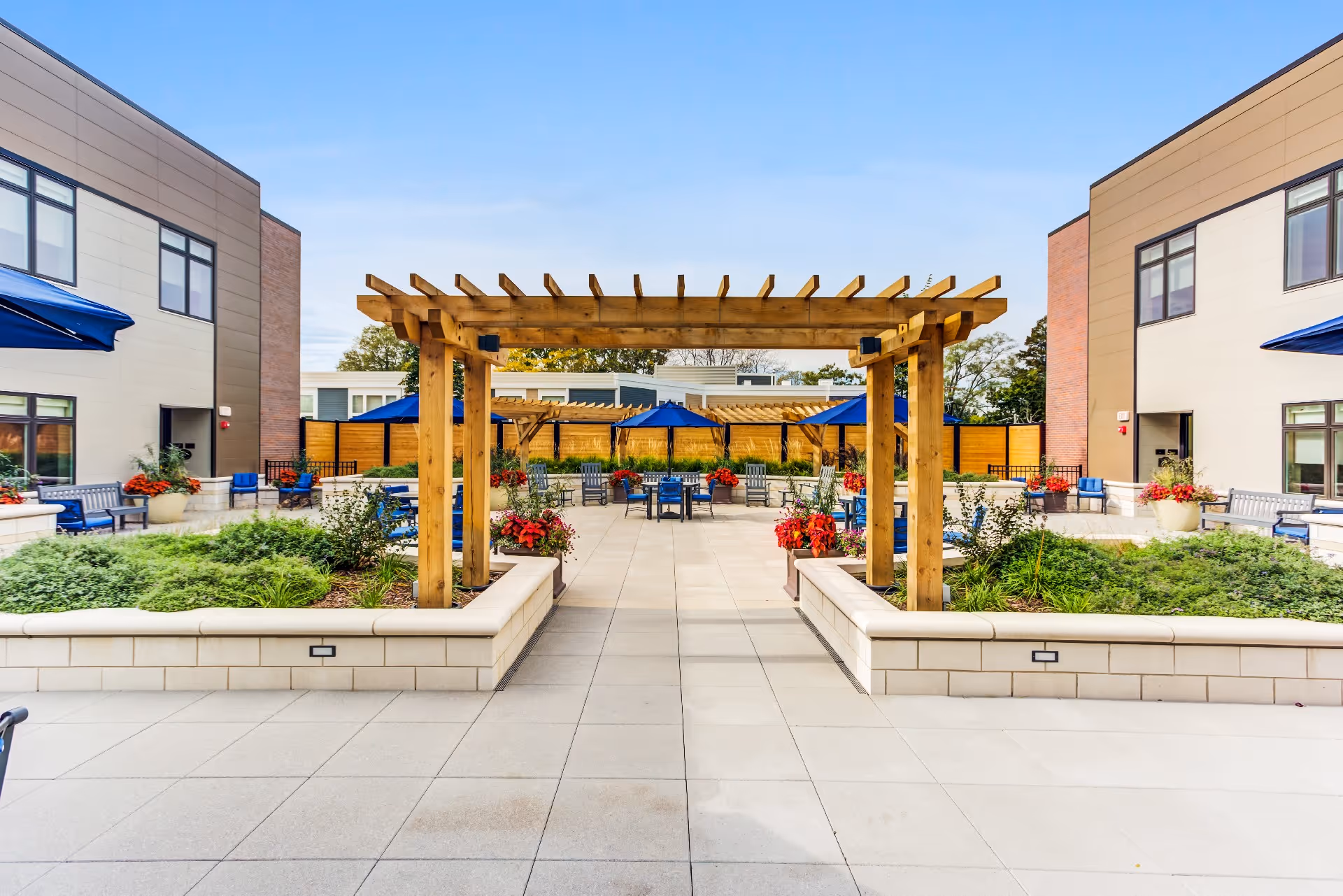Outdoor courtyard of a senior living facility with a central wooden pergola, raised planting beds, patio seating and blue umbrellas between two building wings.