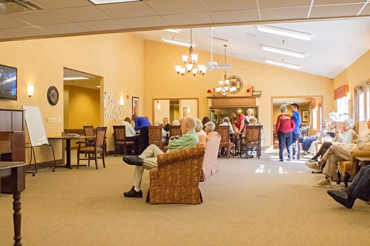A spacious senior living common area with several elderly people seated and interacting. There are chairs and tables arranged around the room, warm yellow walls, ceiling fans, and chandeliers providing lighting. Some people are seated on armchairs, while others are gathered around tables or standing and conversing near the windows.