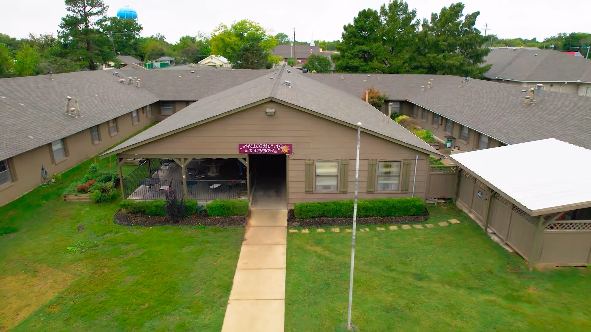 Aerial view of a single-story building complex with a central covered patio area. The building is brown with green shutters and a sign above the entrance that reads 'Welcome to Rainbow'. There is a concrete walkway leading to the entrance, surrounded by green grass and some landscaping. Trees and other buildings are visible in the background.
