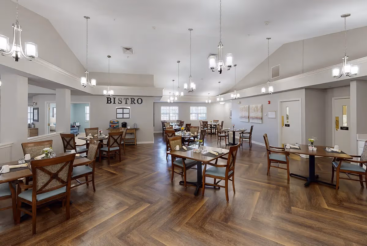 A spacious dining room with wooden floors and multiple tables set with napkins and small flower vases. The room has high ceilings with several modern chandeliers hanging down. On the far wall, the word 'BISTRO' is displayed above a small table with a coffee machine and other items. The walls are painted light gray, and there are windows allowing natural light into the room.