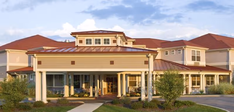 Exterior view of a senior living facility building with a covered entrance supported by white columns, red roof, and beige walls. There are shrubs and a walkway leading to the entrance under a partly cloudy sky.