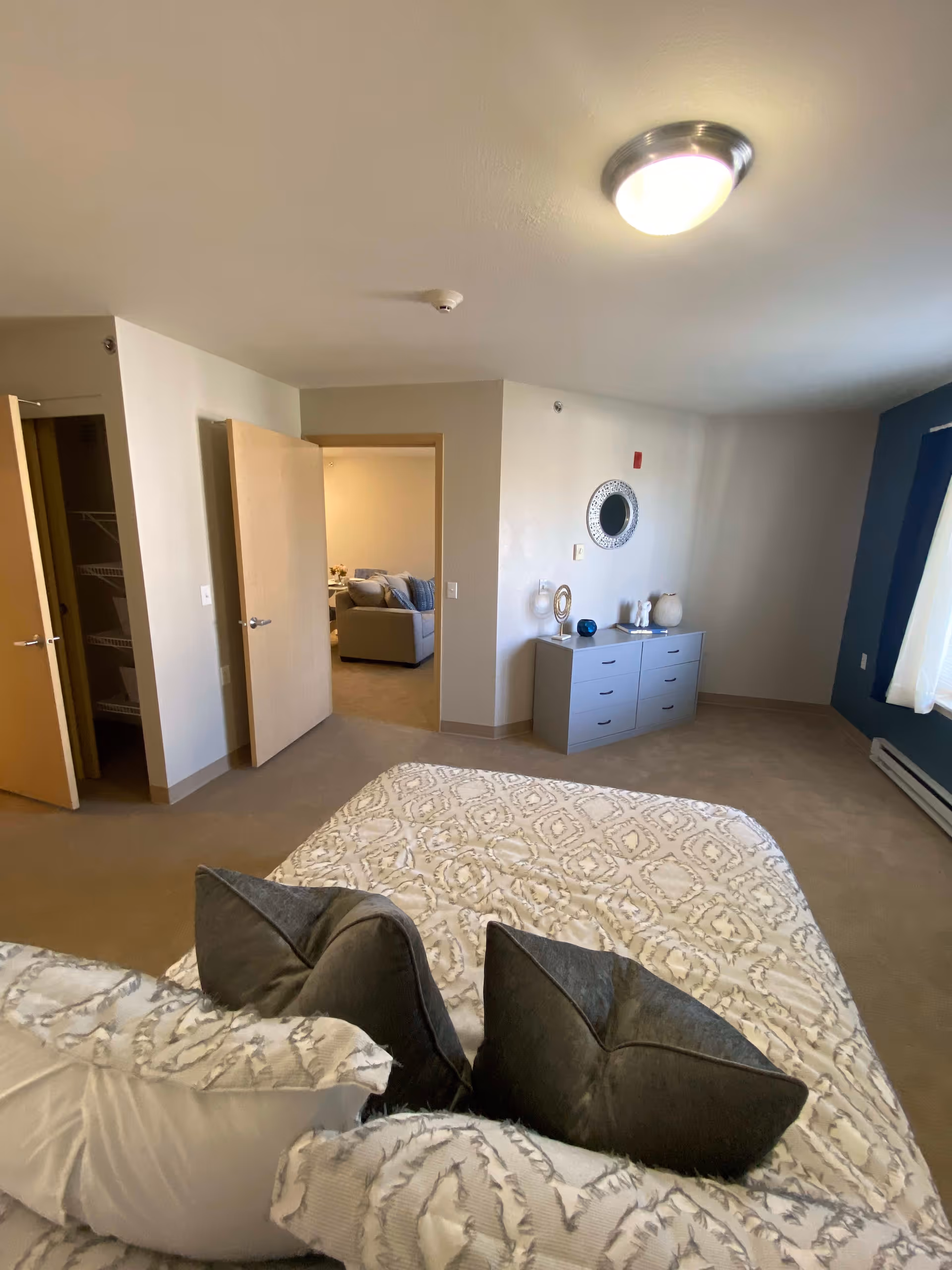 A bedroom in a senior living facility with a bed covered in a patterned beige and white comforter and several pillows. There is a gray dresser against the wall with decorative items and a round mirror above it. The room has beige walls, a window with white curtains, and an open door leading to a living area with a sofa visible.