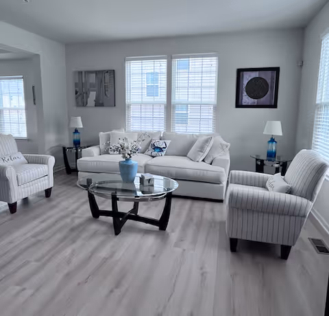 A bright and modern living room with light wood flooring, a white sofa with decorative pillows, two striped armchairs with 'Relax' pillows, a glass coffee table with a blue vase and flowers, two side tables with blue glass lamps, and framed artwork on the walls. Large windows with white blinds allow natural light to fill the room.