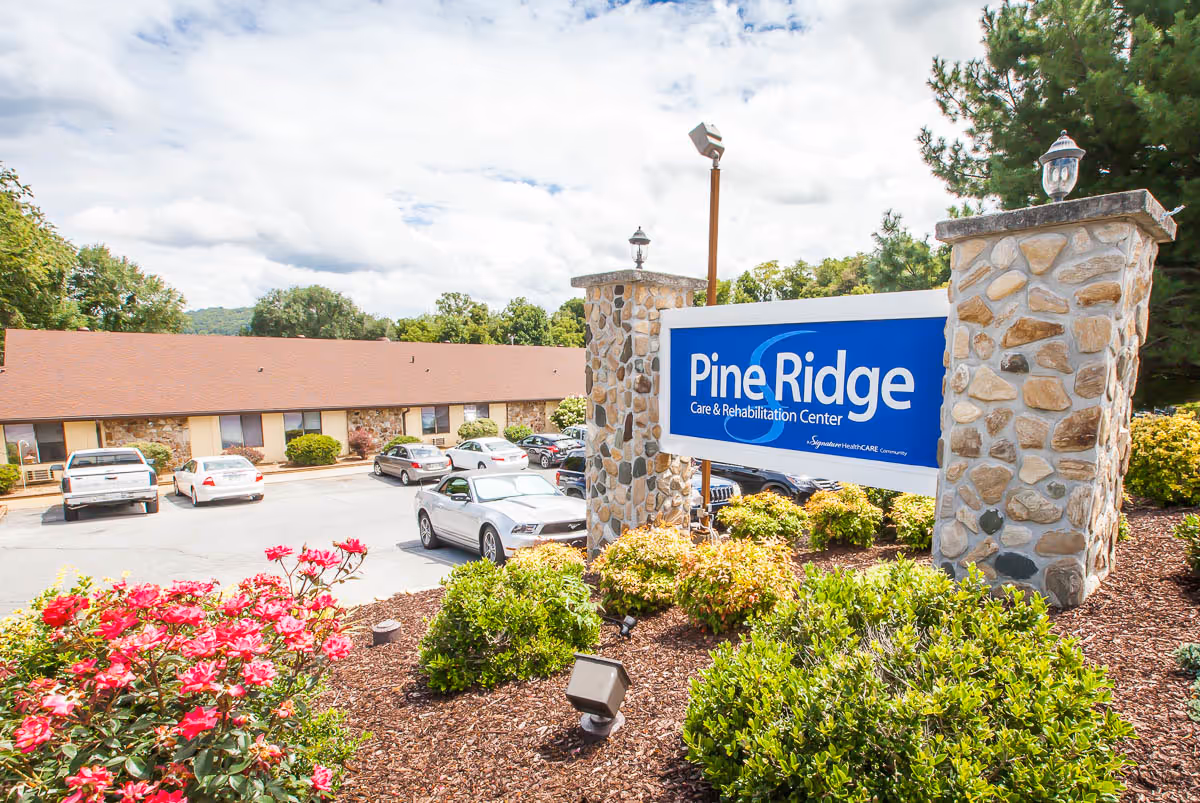 Stone-pillared sign reading "Pine Ridge Care & Rehabilitation Center" in front of a single-story rehab building with parked cars and landscaped shrubs.