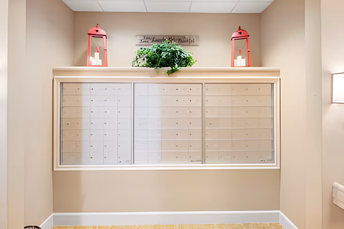Interior view of a beige-colored mailroom with a large set of mailboxes behind a glass window. On top of the window frame, there are two red lanterns with white candles inside and a green leafy plant in the center. A decorative wooden sign is mounted on the wall above the plant.