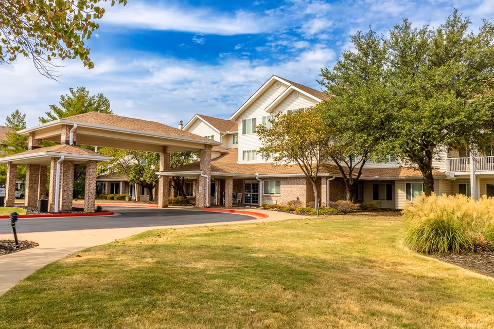 Exterior view of Asher Point Independent Living of Bartlesville showing a multi-story building with a covered entrance driveway, surrounded by trees, grass, and landscaping under a partly cloudy sky.