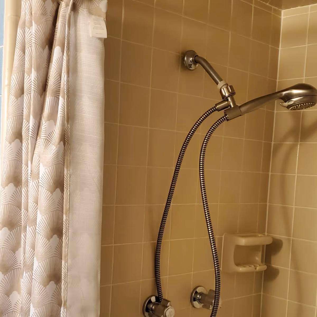 Shower stall with beige tiled walls, a handheld showerhead on a flexible hose and a patterned fabric shower curtain.