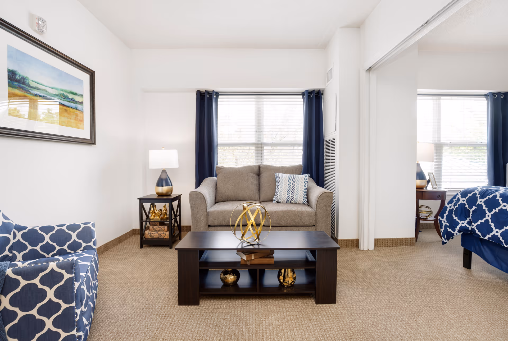 A cozy living room area with a beige loveseat, a dark wooden coffee table with decorative items, a blue and white patterned armchair, and a side table with a lamp. There is a large window with navy blue curtains behind the loveseat. To the right, a bedroom with a blue and white patterned bedspread is partially visible through an open doorway.