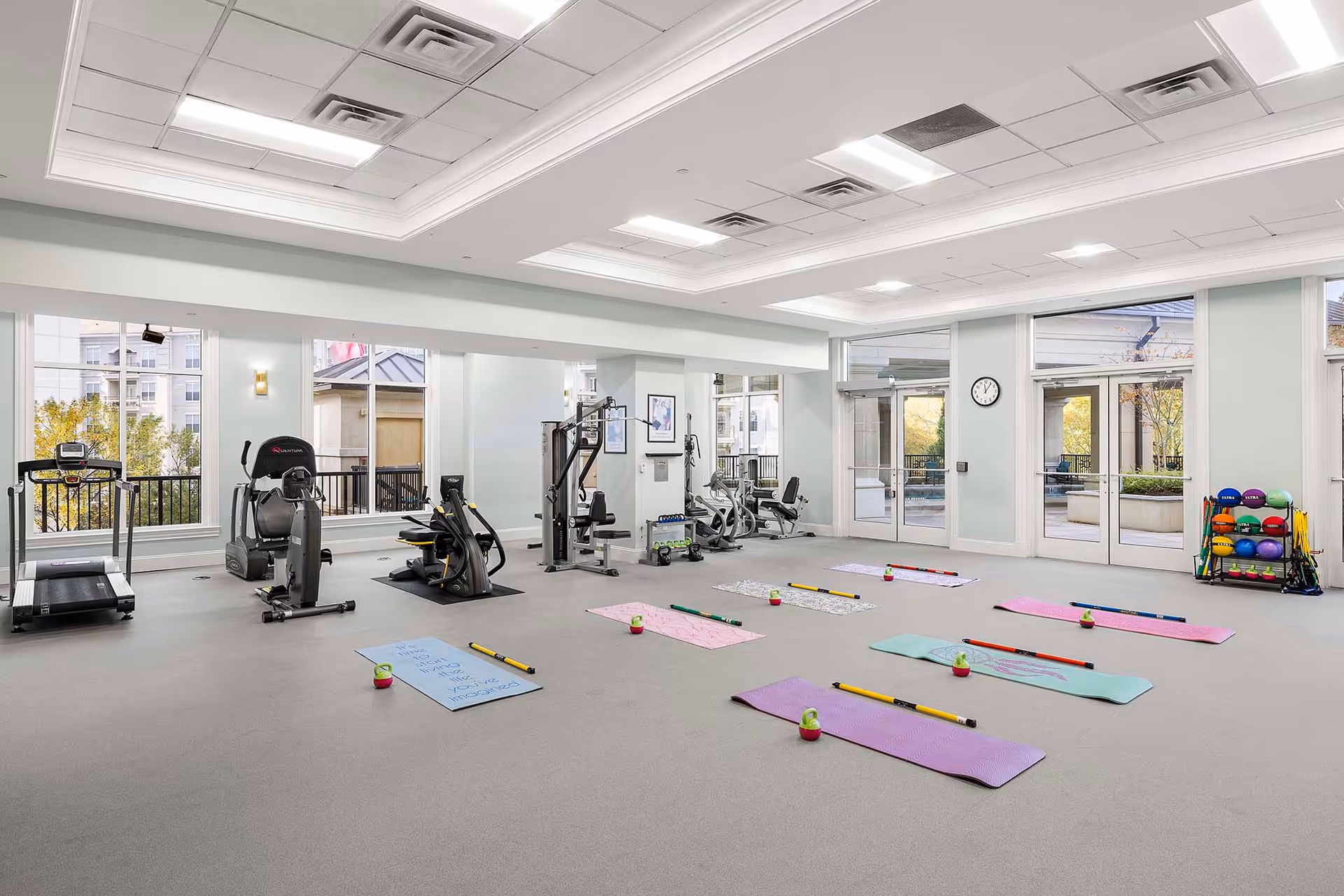 A spacious fitness room with exercise equipment including a treadmill, elliptical machine, and weight machines. Several colorful yoga mats are laid out on the floor, each with a small exercise ball and a weighted bar. Large windows and glass doors let in natural light and provide a view of an outdoor patio area.
