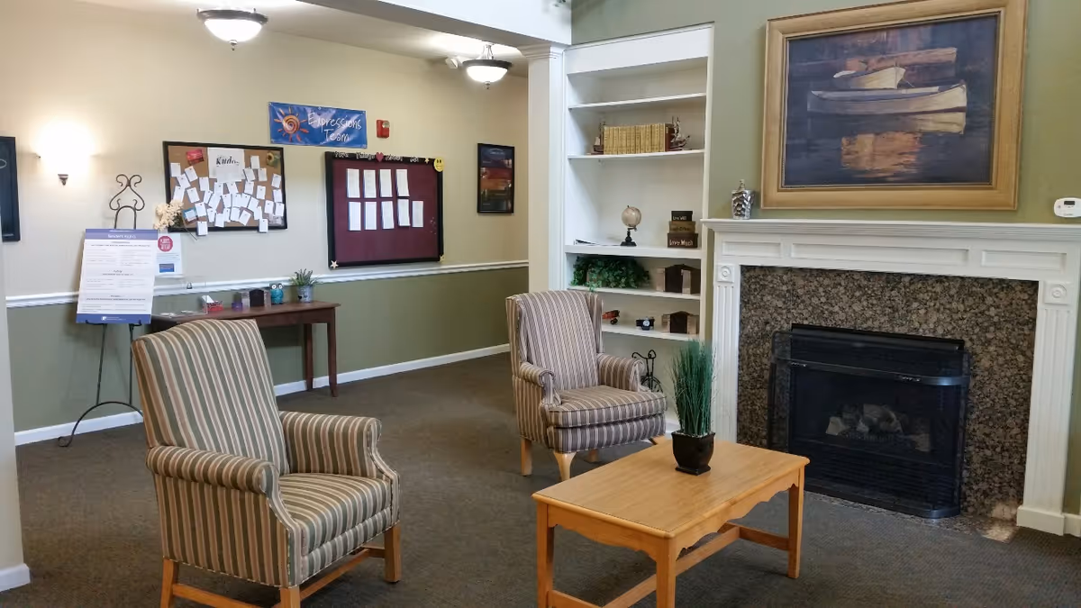Cozy common room with two striped armchairs facing a wooden coffee table, a fireplace, built-in shelves, and bulletin boards on the wall.